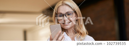 Online Call. Beautiful senior woman in casual wear smiling and talking while making a video call using her smartphone, standing at home 102739535