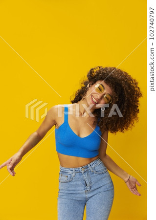 Woman with curly afro hair in a blue T-shirt on a yellow background dancing flying hair with sunglasses yellow, hand signs, look into the camera, smile with teeth and happiness, copy space 102740797
