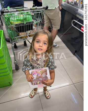 Little girl with a book in her hands stands at the supermarket checkout and looks up Little girl with a book in her hands stands at the supermarket checkout and looks up 102741491