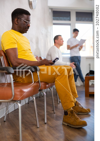 Positive man communicates on mobile phone while sitting on chair in the company office waiting for specialist 102748082