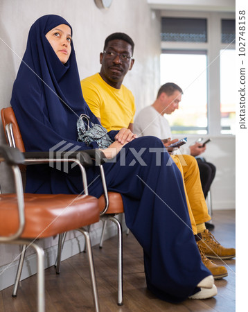 Girl in traditional veil, along with other people of different nationalities, is waiting for a specialist sitting on a chair in hall Girl in traditional veil, along with other people of different nationalities, is waiting for a specialist sitting on a chair in hall 102748158