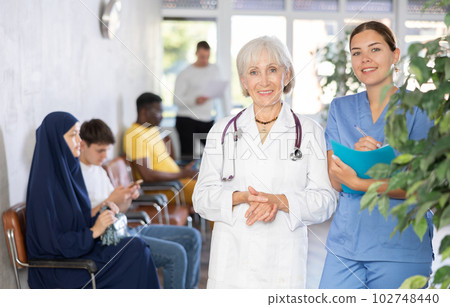 Senior woman standing with young female assistant in clinic lobby 102748440
