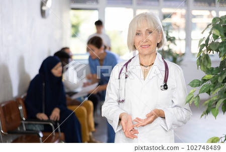 Elderly female doctor standing in busy clinic lobby Elderly female doctor standing in busy clinic lobby 102748489