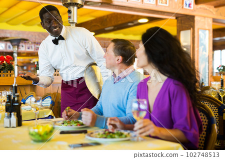 Positive biracial couple talking to friendly African American waiter in restaurant Positive biracial couple talking to friendly African American waiter in restaurant 102748513