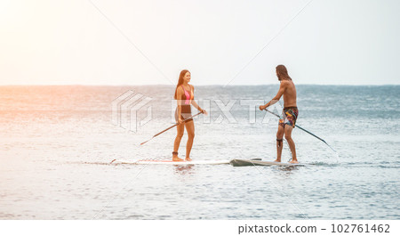 Sea woman and man on sup. Silhouette of happy young woman and man, surfing on SUP board, confident paddling through water surface. Idyllic sunset. Active lifestyle at sea or river. Sea woman and man on sup. Silhouette of happy young woman and man, surfing on SUP board, confident paddling through water surface. Idyllic sunset. Active lifestyle at sea or river. 102761462