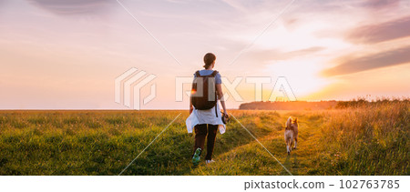 Scenic View Bright Sunbeams. Young Woman Backpacker Walking With Dog In Summer Meadow Grass During Sunset Sunrise Time. Healthy Lifestyle Walks In Evening Summer Sunlight. Panoramic View 102763785