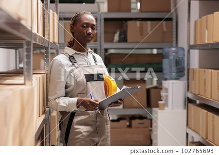 Serious delivery service worker tracking inventory in warehouse portrait. African american shipping business employee checking supply, holding clipboard and writing in checklist Serious delivery service worker tracking inventory in warehouse portrait. African american shipping business employee checking supply, holding clipboard and writing in checklist 102765693