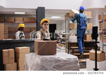 Young woman explaining warehouse worker job at mobile phone camera. Industrial storehouse operator talking about parcel receiving and dispatching management at smartphone Young woman explaining warehouse worker job at mobile phone camera. Industrial storehouse operator talking about parcel receiving and dispatching management at smartphone 102765754