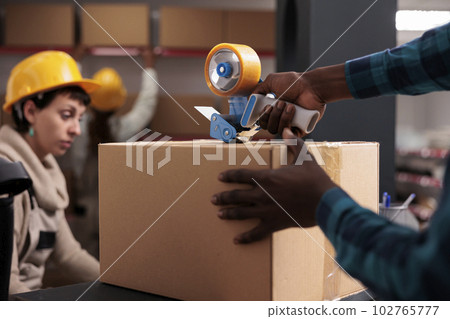 Delivery office manager packing parcel using hand held sticky tape dispenser. African american shipping department worker arms closing cardboard box with adhesive close up 102765777
