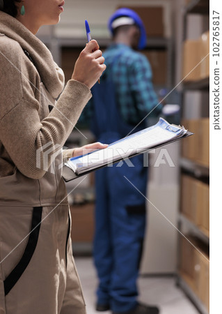 Industrial warehouse employees managing goods and working in stockroom. Storehouse operative woman wearing uniform overall counting parcels cardboard boxes and writing on clipboard Industrial warehouse employees managing goods and working in stockroom. Storehouse operative woman wearing uniform overall counting parcels cardboard boxes and writing on clipboard 102765817