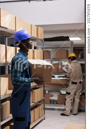 Mail sorting center worker holding packed cardboard box with client order. Shipping company african american man employee carrying carton and managing parcels transportation Mail sorting center worker holding packed cardboard box with client order. Shipping company african american man employee carrying carton and managing parcels transportation 102765859