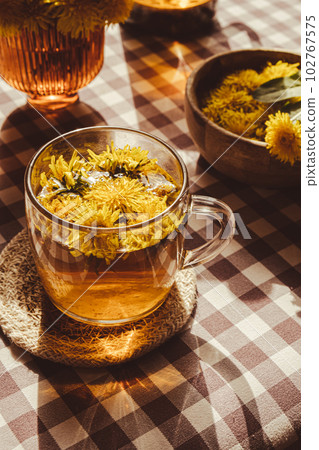 Dandelion flower healthy tea in glass teapot and glass cup on table. Delicious herbal Hot tea from fresh dandelion flowers at home at summer day. Green clearing. Bouquet of dandelions petals Dandelion flower healthy tea in glass teapot and glass cup on table. Delicious herbal Hot tea from fresh dandelion flowers at home at summer day. Green clearing. Bouquet of dandelions petals 102767575
