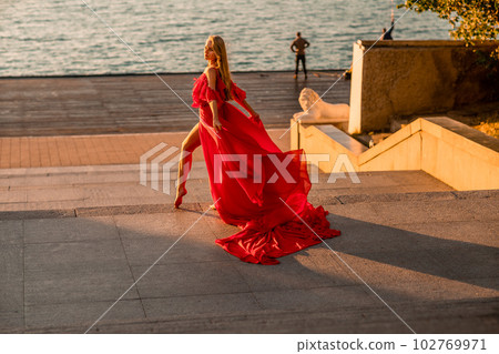 Sunrise red dress. A woman in a long red dress against the backdrop of sunrise, bright golden light of the sun's rays. The concept of femininity, harmony. 102769971