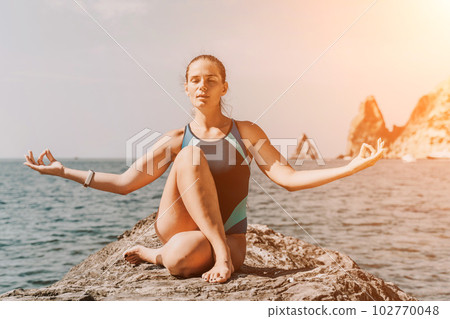 Yoga on the beach. A happy woman meditating in a yoga pose on the beach, surrounded by the ocean and rock mountains, promoting a healthy lifestyle outdoors in nature, and inspiring fitness concept. 102770048