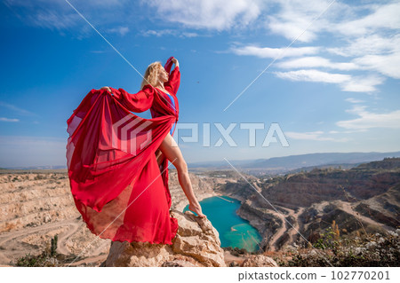 woman red dress lake mountains. Side view of a woman in a long red dress posing on a rock high above the lake. Against the background of the blue sky and the lake in the form of a heart 102770201