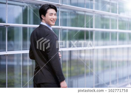 一位身著黑髮西裝的男子微笑著走在辦公樓前 複製空間可用 102771758