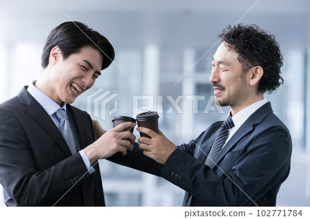 A boss in a suit and a subordinate office worker talking while toasting with coffee. Smiling upper body, close up. A boss in a suit and a subordinate office worker talking while toasting with coffee. Smiling upper body, close up. 102771784