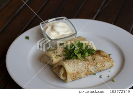 Two halves of pancake roll with raspberry filling, sour cream in white plate, teaspoon in bowl with sour cream, fork on wooden table. Two halves of pancake roll with raspberry filling, sour cream in white plate, teaspoon in bowl with sour cream, fork on wooden table. 102772549