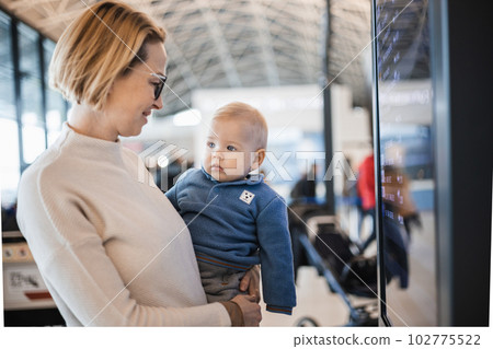 Mother traveling with child, holding his infant baby boy at airport terminal, checking flight schedule, waiting to board a plane. Travel with kids concept Mother traveling with child, holding his infant baby boy at airport terminal, checking flight schedule, waiting to board a plane. Travel with kids concept 102775522
