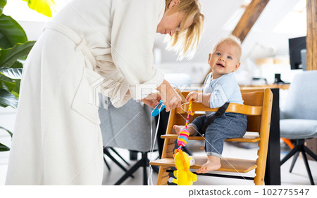 Happy infant sitting and playing with his toy in traditional scandinavian designer wooden high chair in modern bright atic home suppervised by his mother Happy infant sitting and playing with his toy in traditional scandinavian designer wooden high chair in modern bright atic home suppervised by his mother 102775547