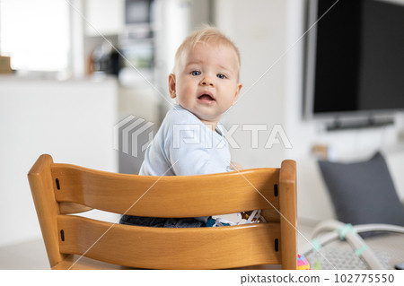 Happy infant sitting in traditional scandinavian designer wooden high chair and laughing out loud in modern bright home. Cute baby smile Happy infant sitting in traditional scandinavian designer wooden high chair and laughing out loud in modern bright home. Cute baby smile 102775550