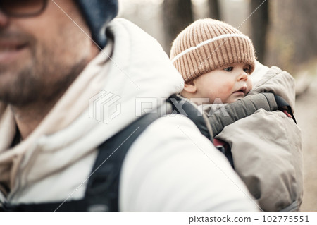 Sporty father carrying his infant son wearing winter jumpsuit and cap in backpack carrier hiking in autumn forest Sporty father carrying his infant son wearing winter jumpsuit and cap in backpack carrier hiking in autumn forest 102775551