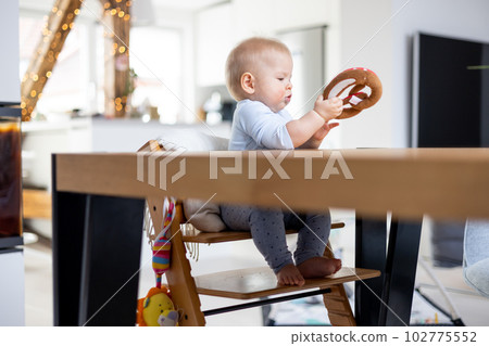 Happy infant sitting at dining table and playing with his toy in traditional scandinavian designer wooden high chair in modern bright atic home. Cute baby playing with toys. Happy infant sitting at dining table and playing with his toy in traditional scandinavian designer wooden high chair in modern bright atic home. Cute baby playing with toys. 102775552