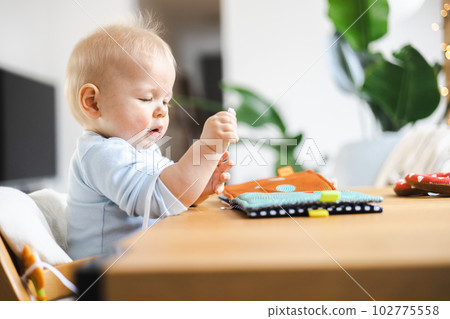Happy infant sitting at dining table and playing with his toy in traditional scandinavian designer wooden high chair in modern bright atic home. Cute baby playing with toys. Happy infant sitting at dining table and playing with his toy in traditional scandinavian designer wooden high chair in modern bright atic home. Cute baby playing with toys. 102775558