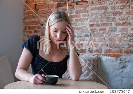 Sad tired young woman with a coffee in a cafe touching forehead having headache migraine or 102775947