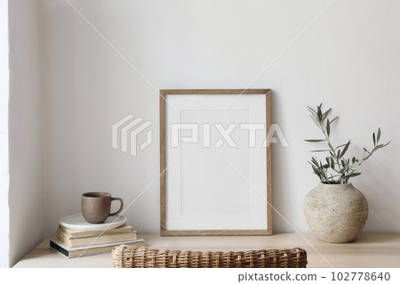 Closeup of picture frame, poster mockup. Vase with olive tree branches on wooden table. Blurred rattan chair. Cup of coffee, books. Summer artistic Mediterranean interior. Working space, home office. 102778640