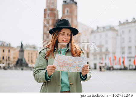 Attractive young female tourist is exploring new city. Redhead girl walking and holding a paper map in Krakow. Traveling Europe in spring. Urban lifestyle banner 102781249