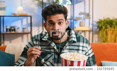 Indian man sits on couch eating popcorn and watching interesting TV serial sport game online at home 102785695