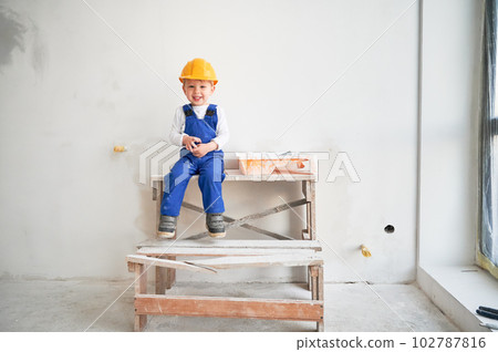 Cute kid construction worker sitting on wooden table against white wall in apartment under renovation. Cheerful little boy wearing safety helmet and work overalls while looking at camera and laughing. 102787816