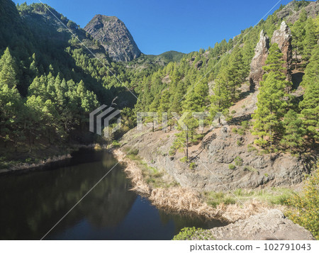 Water reservoir, dam and pine forest at small village La Laja along hiking trail with Los Roques, Roque de Ojila in the background, Garajonay National Park, La Gomera, Spain Water reservoir, dam and pine forest at small village La Laja along hiking trail with Los Roques, Roque de Ojila in the background, Garajonay National Park, La Gomera, Spain 102791043
