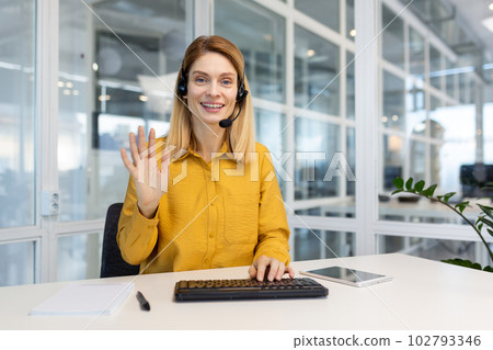 Portrait of a young woman in a headset who sits in the office at the keyboard and greets the camera with her hand. Works in support and assistance service, telemarket, call center. 102793346