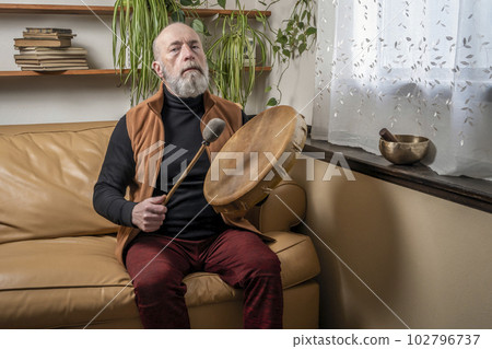senior man is playing a native American style, shaman drum with a beater at home, lifestyle and hobby concept 102796737