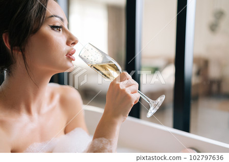 Closeup face of glamour young woman with beautiful hairstyle and makeup drinking champagne sitting in foam bath enjoying alcoholic drink. Pretty brunette female relaxing in bathroom at home. 102797636