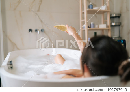 Back view of unrecognizable sexy naked young woman pouring out champagne in bathtub at home. Closeup of beautiful brunette female relaxing in bath in luxury bathroom with alcohol, back view. Back view of unrecognizable sexy naked young woman pouring out champagne in bathtub at home. Closeup of beautiful brunette female relaxing in bath in luxury bathroom with alcohol, back view. 102797643