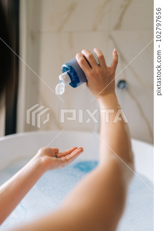 Vertical back view of unrecognizable young woman taking bubble bath, pouring shampoo into hand, enjoying home spa procedure, selective focus. Lovely millennial lady pampering skin, lying in bathtub. Vertical back view of unrecognizable young woman taking bubble bath, pouring shampoo into hand, enjoying home spa procedure, selective focus. Lovely millennial lady pampering skin, lying in bathtub. 102797656