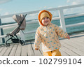 A toddler in a yellow jacket and pants walks on the pier 102799104