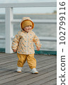 A toddler in a yellow jacket and pants walks on the pier 102799116