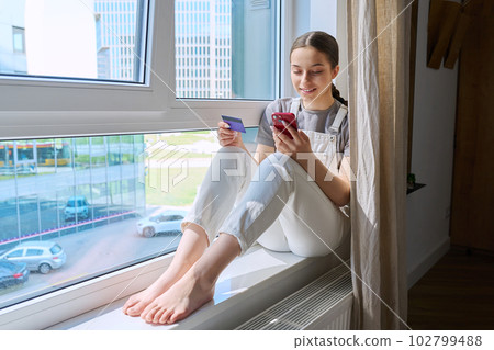 Teenage girl with smartphone and credit card sitting on windowsill at home 102799488