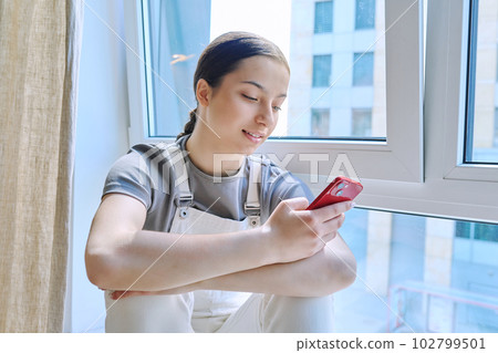 Teenage girl sitting at home near window with smartphone Teenage girl sitting at home near window with smartphone 102799501