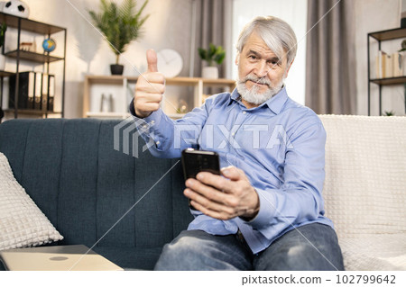 Gray-haired man using phone while taking seat in living room Gray-haired man using phone while taking seat in living room 102799642