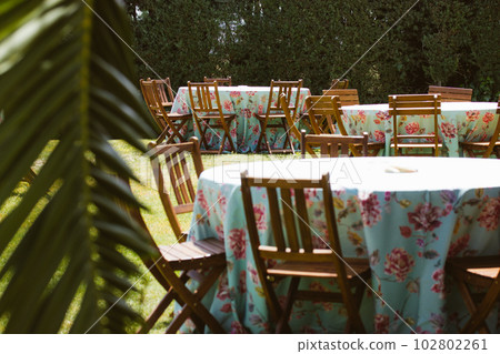 Beautifully set tables on backyard. Private party outdoors preparation. Comunion in Spain, traditional catholic holiday celebration. Garden furniture, wooden tables and chairs among palm tree leaves. 102802261