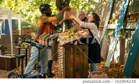 Happy man with bike carrying backpack to deliver food from farmers market stand, picking up box of bio produce. Man courier delivering fruits and vegetables produced by local farmers to customers. 102804239