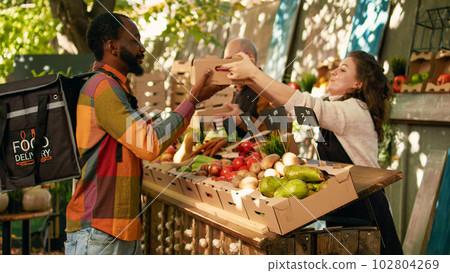 African american deliveryman wearing backpack waiting for order at farmers market counter. Team of farmers delivering fresh organic fruits and vegetables to customers, small business owners. African american deliveryman wearing backpack waiting for order at farmers market counter. Team of farmers delivering fresh organic fruits and vegetables to customers, small business owners. 102804269