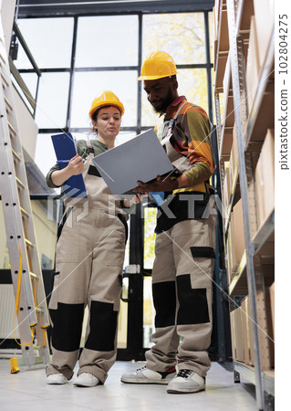 Multi ethnic team looking at merchandise checklist, working at goods inventory in storehouse. African american workers holding laptop computer analyzing online orders preparing packages for delivery Multi ethnic team looking at merchandise checklist, working at goods inventory in storehouse. African american workers holding laptop computer analyzing online orders preparing packages for delivery 102804275