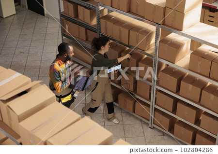 Warehouse manager scanning carton boxes, making merchandise inventory on digital tablet. Shipping service company team in protective overalls preparing orders, working in storehouse. Top view Warehouse manager scanning carton boxes, making merchandise inventory on digital tablet. Shipping service company team in protective overalls preparing orders, working in storehouse. Top view 102804286