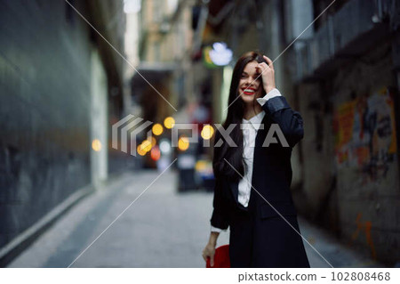 Fashion woman smile with teeth and fun tourist in stylish clothes in jacket and red beret walking down narrow city street flying hair, travel, French style, cinematic color, retro style. Fashion woman smile with teeth and fun tourist in stylish clothes in jacket and red beret walking down narrow city street flying hair, travel, French style, cinematic color, retro style. 102808468